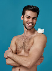 Fototapeta premium Studio shot of joyful naked man posing with white jar of effective cream on his shoulder, smiling at camera, standing with arms crossed isolated over blue background