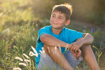 happy teenage boy in blue t-shirt sits on the grass, dreaming, in backlight