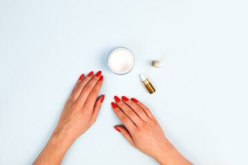 Top view of female hands with red nails, open cream jar and glass bottles with pipette on blue table. Skin care, moisturizing beauty products, lifestyle. Concept hand care