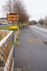 Obraz premium Information electric sign with message expect delays by a small road. House estate on the left. Selective focus, cloudy sky. Low car traffic on the road.