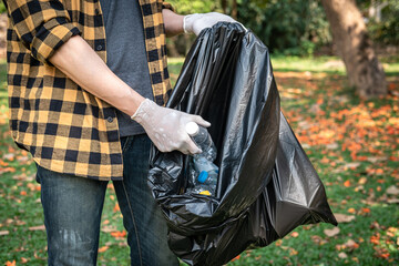 Volunteer man in gloves to picking up plastic bottle into plastic black bag for cleaning the park