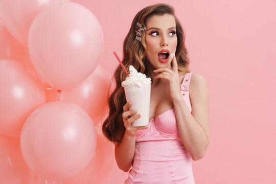 Surprised woman eating cream while posing with balloons and milkshake