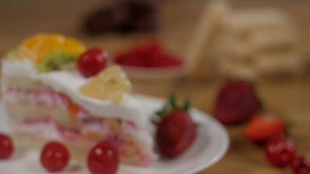 Camera focus on a slice of creamy sponge cake decorated with fresh fruits. Closeup shot of a red ripe juicy strawberry and cherries served with a pastry placed on a plate