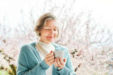 Spring portrait of beautiful and elegant middle age woman posing against pink blooming tree, holding cup of coffee or tea