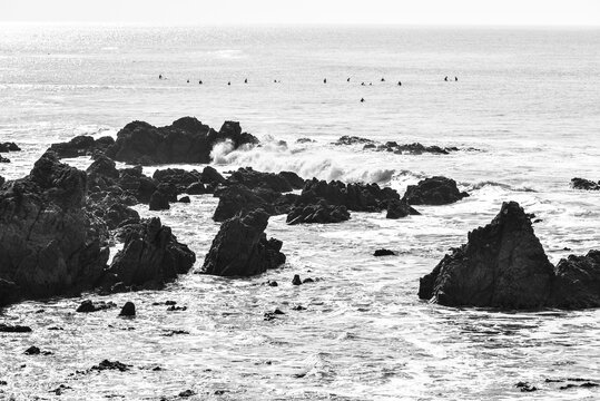 Black and white photo of the ocean and rocks, with surfers in the background waiting for a wave.