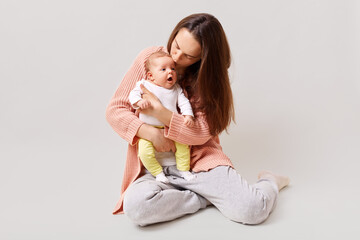 Obraz na płótnie Canvas Young beautiful attractive mother holding and kissing newborn baby while sitting on floor, dark haired female wearing casual attire, isolated over white background.