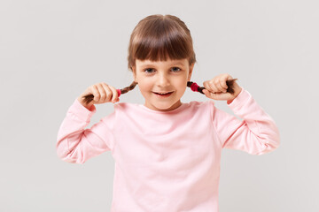Little cute preschool girl looks at the camera with smile, dark-haired female child holding pigtails in her hands, wearing a casual jumper, isolated over white background.