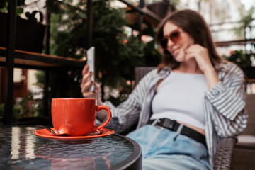 Young Cheerful Woman Using Smartphone While Sitting in Cafe Outdoors, Focus on Cup of Coffee