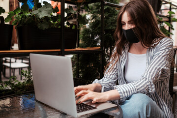 Young Brunette Woman Wearing Protective Face Mask Working on Laptop While Sitting in Cafe Outdoors, Pandemic Covid-19 Concept