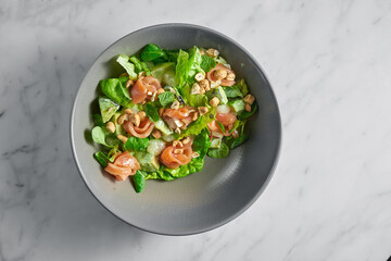 Fresh salad with salmon, delicate cheese, peanuts and mint in a gray bowl on a white marble background