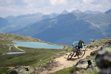 Girl with a mountainbike on a single trail in front of peaks mountains lake and rocks in the austrian alps
