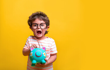 Portrait of little funny boy putting money on a moneybox. Kid in glasses isolated on yellow....