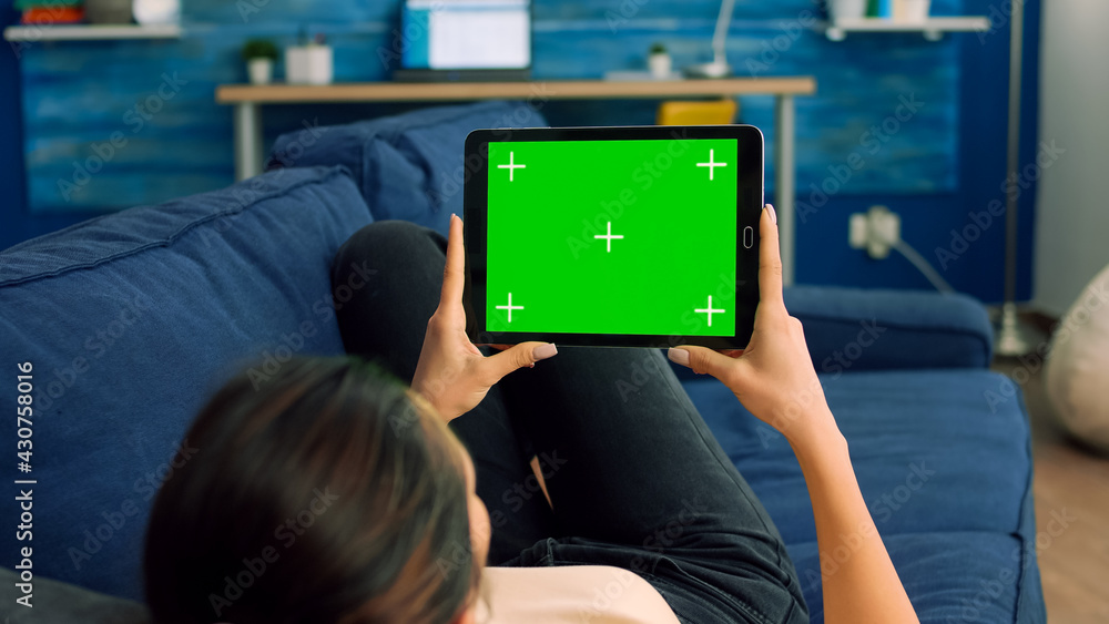 Poster over the shoulder view of casual woman sitting on sofa using tablet computer with mock up green scre
