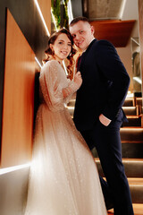 Naklejka premium The groom in a suit and the bride in a white dress stand on poorly lit staircase