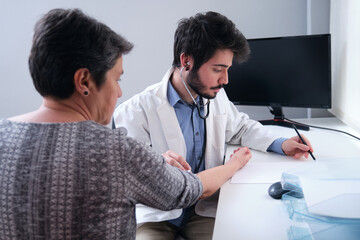 Young doctor listening to mature woman patient heart with a stethoscope and writing down. Medical exam.