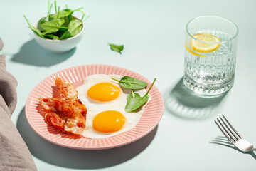 English breakfast - eggs, bacon and spinach leaves in a plate. Water with lemon. Hard light.
