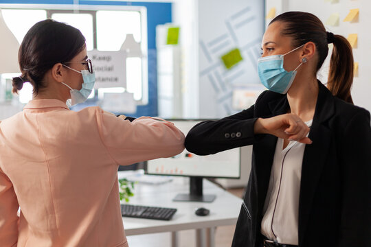 Woman In Business Office Following Safety Precaution Wearing Face Mask, Touching Elbow During Global Pandemic With Covid19 Flu. Colleaues Greeting Eachother.