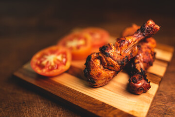 Close of roasted chicken drumsticks decorated on wooden plate along with fresh, juicy tomatoes. Dramatic image of non vegetarian food with copy space for text.