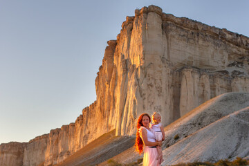 beautiful happy girl with long red hair holds baby in her arms, behind them huge impressive rock sunset