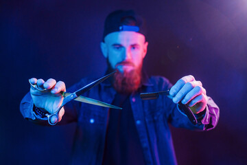 Young bearded barber in cap standing in the studio with neon lighting