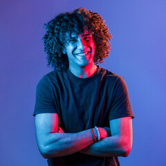 Posing for a camera. Young beautiful man with curly hair is indoors in the studio with neon lighting - obrazy, fototapety, plakaty