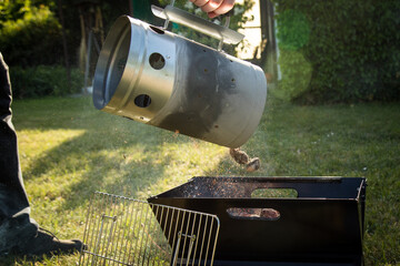 Mann gibt glühende Kohle von Anzündkamin in Klappgrill nach Anheizen für Glut zum grillen draußen im Garten 