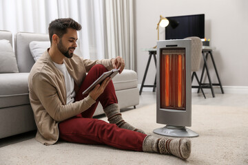 Young man reading book near electric heater at home