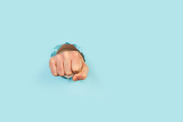 Mano de hombre con el puño cerrado dando un golpe y perforando y rompiendo un papel celeste. Vista de frente y de cerca. Copy space