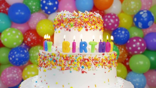 Cake with Happy Birthday candles. Seamless loop. Three-tier butter cream pie with candy sprinkles on rotating cake stand, colorful balloons in background