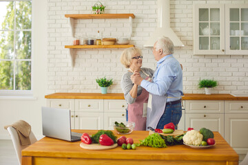 Happy loving mature couple having fun and dancing waltz while cooking healthy vegetarian meal in modern Scandinavian kitchen at home. Active senior citizens spending time together and enjoying life