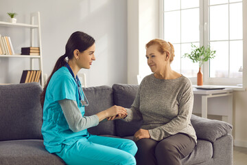 Obraz premium Health consultation for old people. Female nurse supporting an elderly patient holding her hand during a medical visit home. Ambulance worker calms a woman sitting on the couch.