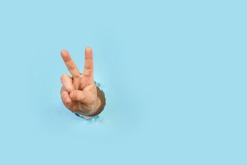 Mano masculina levantada y mostrando un gesto de paz con dos dedos levantados a traves de un agujero en un papel celeste. Vista de frente y de cerca. Copy space