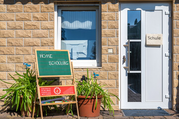 Hauswand mit Tür und Fenster und einer Kreidetafel mit dem Wort Homeschooling und eines Papierzettel an der Tür mit dem Wort Schule in deutscher Sprache