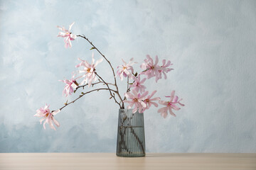 Magnolia tree branches with beautiful flowers in glass vase on wooden table against light blue background