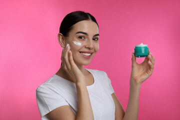 Young woman applying facial cream on pink background