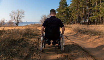A man rides in a wheelchair on the road. A disabled man in a wheelchair rides heavily on a sandy road along the forest. Rear view. Selective focus