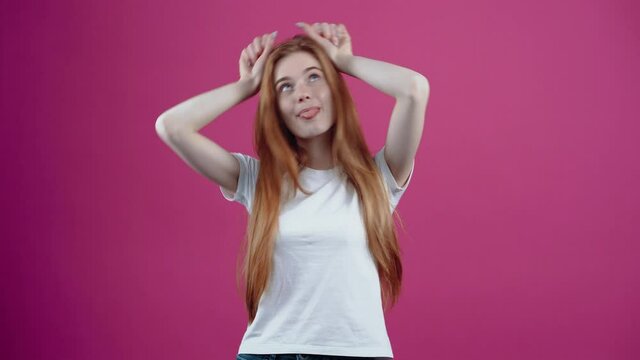 The portrait of a charismatic young woman who playfully puts her hands to her head like bunny ears. Freckled teenage girl in a white T-shirt, isolated on a pink background. The concept of people's