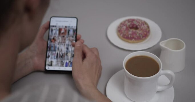 Woman at Home Sitting at Table With Coffee and Donut using Smartphone Scrolling Through Instagram Feed. Girl Looks Social Network  Page On Mobile Phone. Woman Using Social Network on Smartphone.