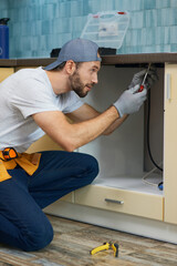 Fixing leaks. Focused young repairman, professional plumber wearing tool belt crouching on the floor while fixing sink pipe in the kitchen indoors