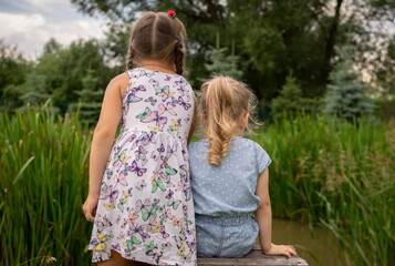 two girls are sitting in a forest park near a pond on a bench