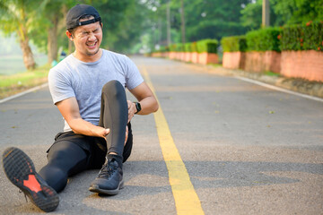 Exercise Injury Concept: A young Asian runner sits down on a park street due to pain in his ankle...
