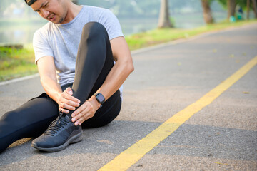 Exercise Injury Concept: A young Asian runner sits down on a park street due to pain in his ankle...
