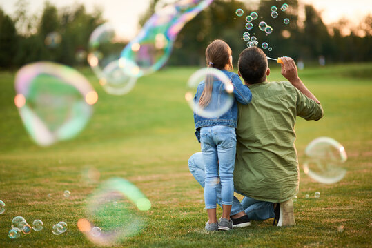 Rear view of young father and his cute little girl having fun while blowing soap bubbles in the park, daughter and dad spending time together outdoors