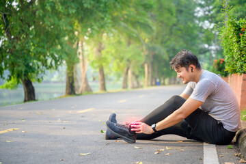 Obraz premium Exercise Injury Concept: A young Asian runner sits down on a park street due to pain in his ankle and grabs his sore ankle and flexes his leg while in severe pain.