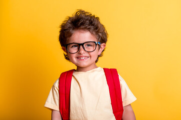 Happy boy in eyeglasses and with backpack isolated on yellow background. Smart little child go to...