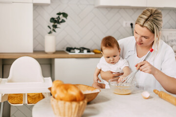 Obraz na płótnie Canvas A young mother holds her baby in her arms, whips the eggs with a whisk and prepares the dough. Mom and baby spend time together