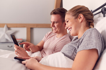 Couple In Bed Reading And Looking At Mobile Phone Together