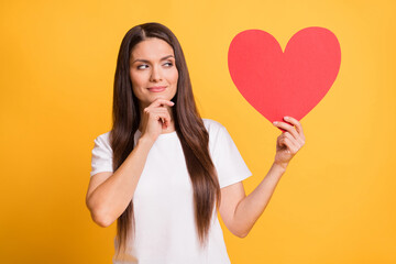 Photo of minded doubtful mature woman look hold big red paper heart wear white t-shirt isolated on yellow color background