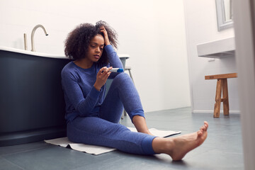 Disappointed Woman Sitting On Floor In Bathroom At Home With Negative Home Pregnancy Test