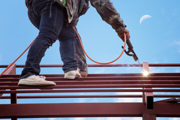 Red painted steel roof truss, with roof welders working at dangerous heights. Welding of steel is very technically demanding and is only professional. Bright sky background